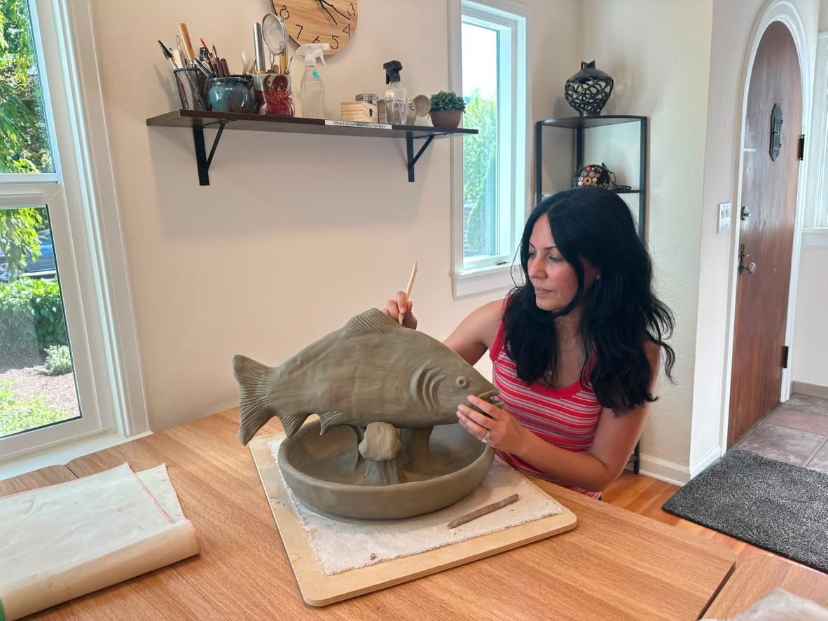 Elmira carving details into a clay fish sculpture at her studio table