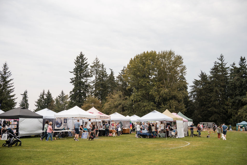 A long row of white 10x10 vendor tents on an open field, ringed by evergreens, with shoppers moving between them