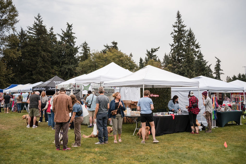 Shoppers and their dogs browsing a row of white vendor tents on the grass at a Making Local market