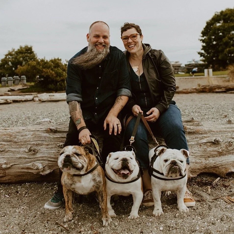 Sarah and her husband Brad sitting on driftwood at the beach with their three bulldogs