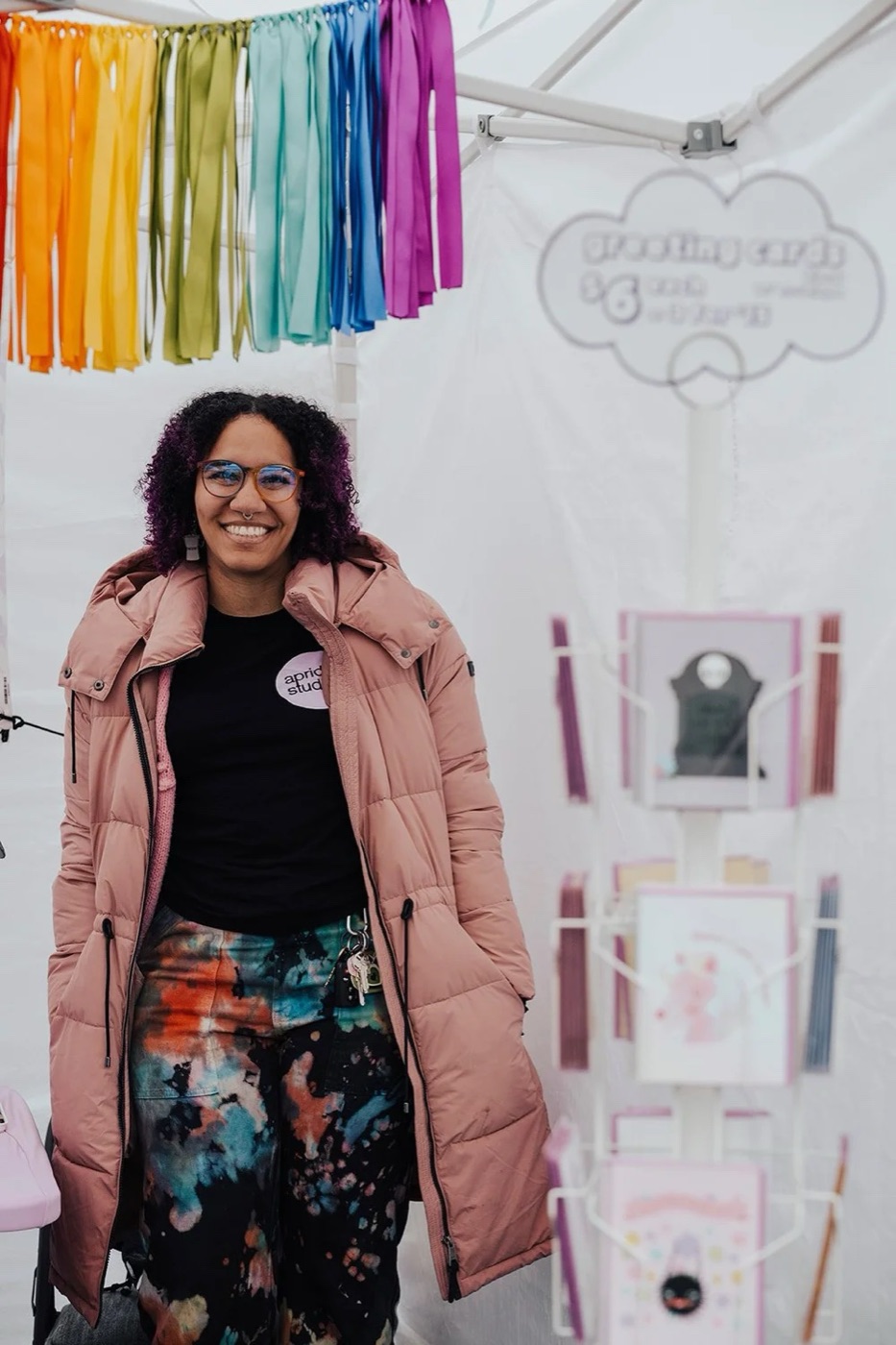 A vendor with purple hair standing in front of a display of rainbow ribbons and greeting cards