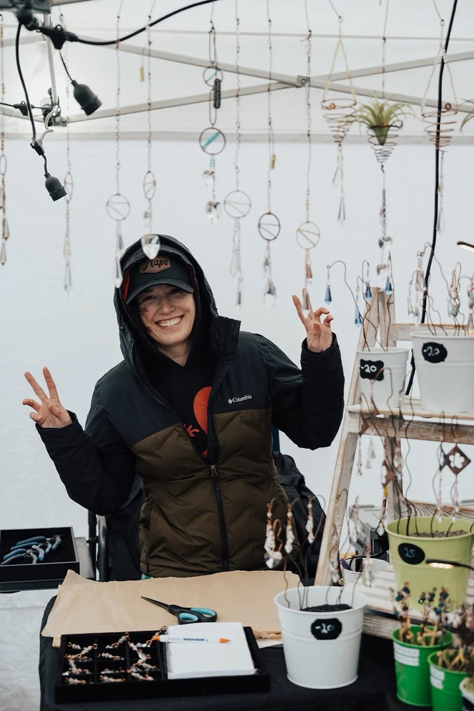 A vendor in a hooded jacket throwing peace signs at her booth of hanging sun catchers and small potted plants